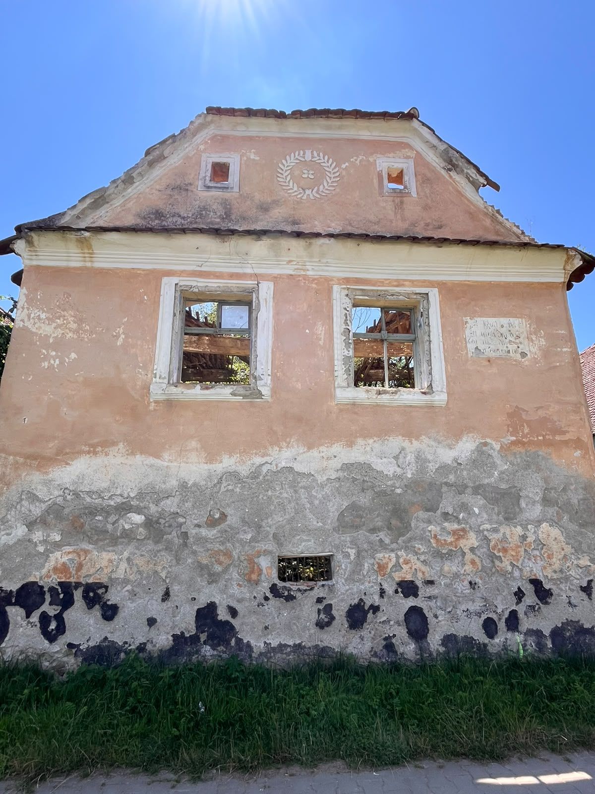 Fațada unei case săsești în ruină din Dacia — fronton cu cunună de lauri, ferestre goale spre cer, acoperiș prăbușit